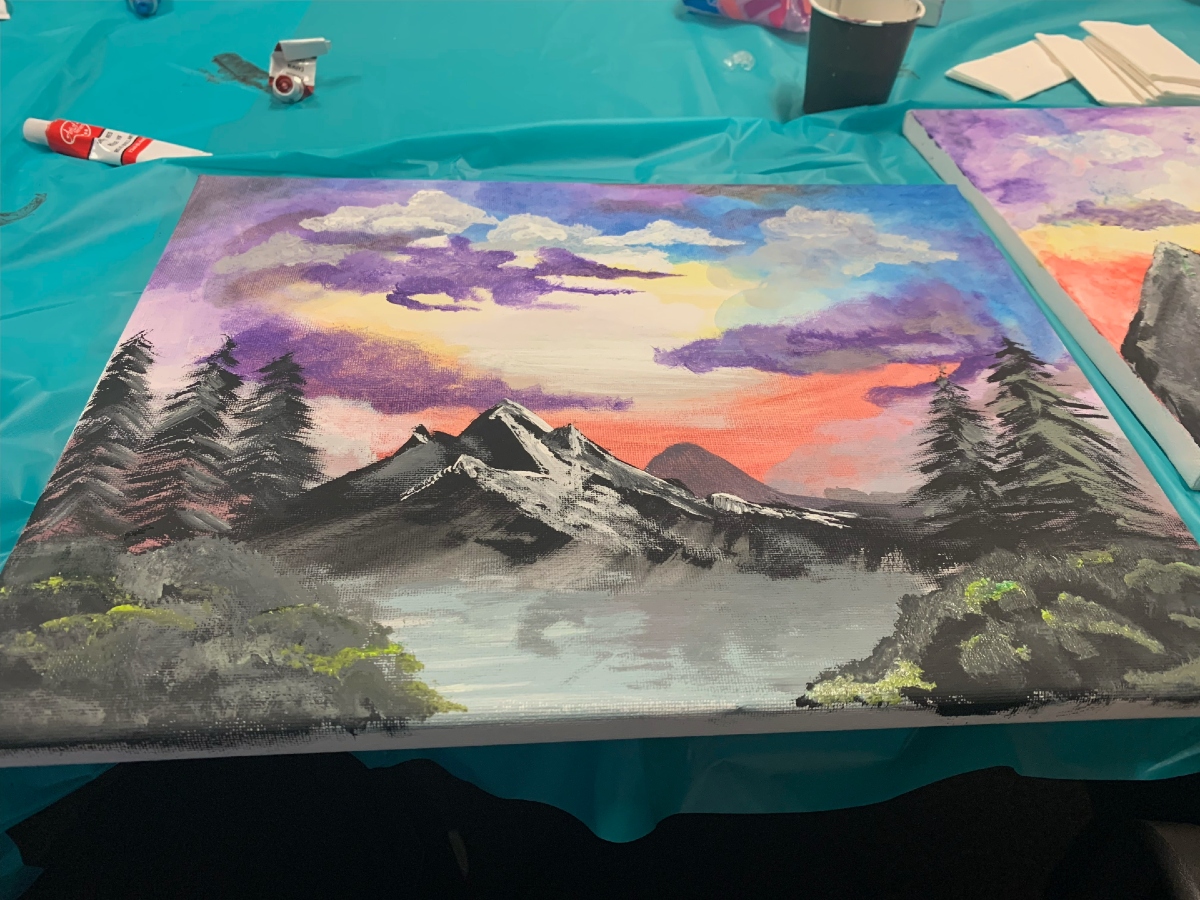 Painting of purple skies and mountains lay on a turquoise table sheet with paint supplies surrounding it.