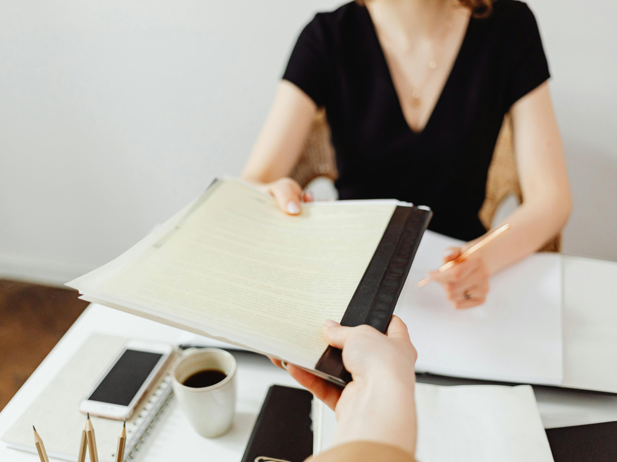 An individual passes a folder to a client over a table with notebooks, a coffee cup and pencils. 