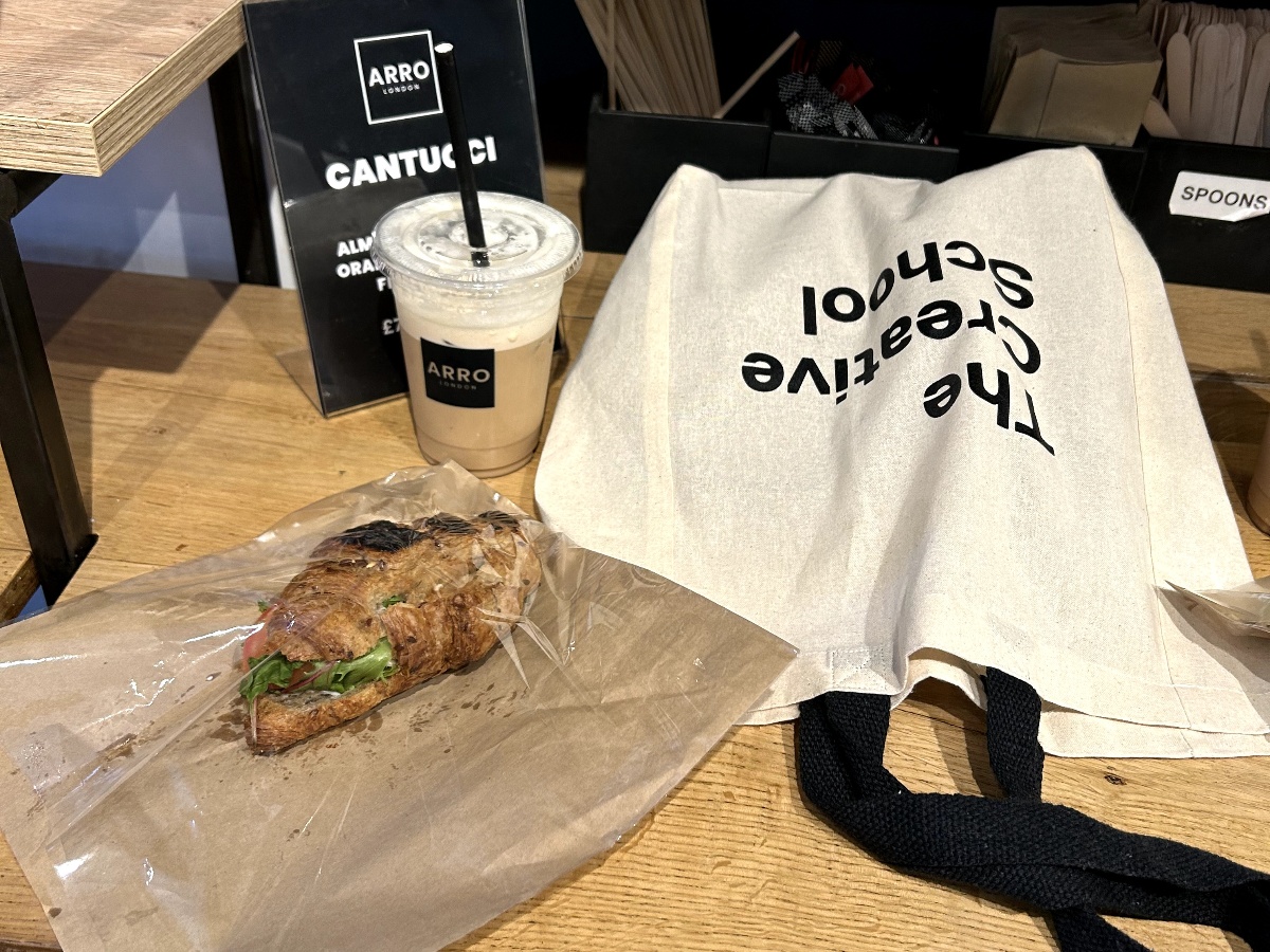 A tote bag from The Creative School, coffee and sandwich lay on a wooden table in the serene Arro café.