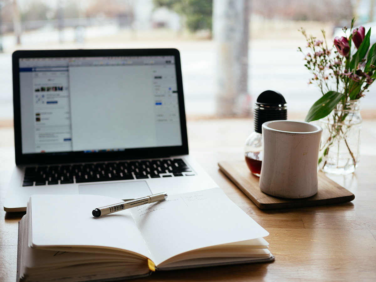 A pen rests on a notebook in front of a laptop on a desk with a cup of coffee on the side. 
