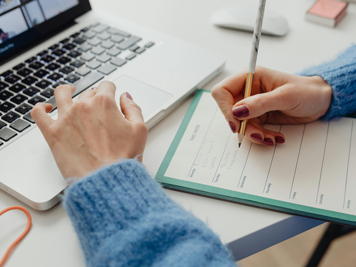 A close-up of a person writing their weekly to-do list on a notepad and scrolling on their laptop.