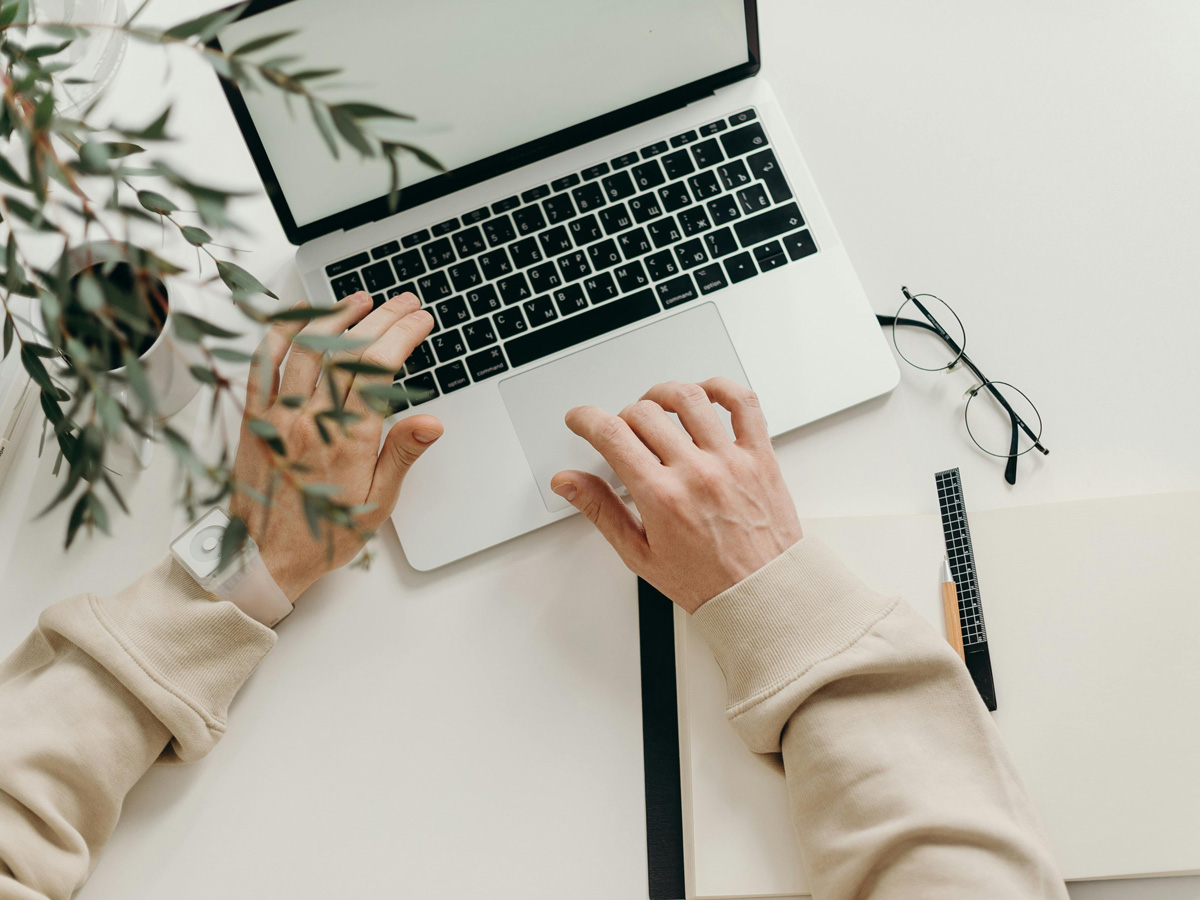 A student typing on their laptop next to an open notebook, glasses and a plant.
