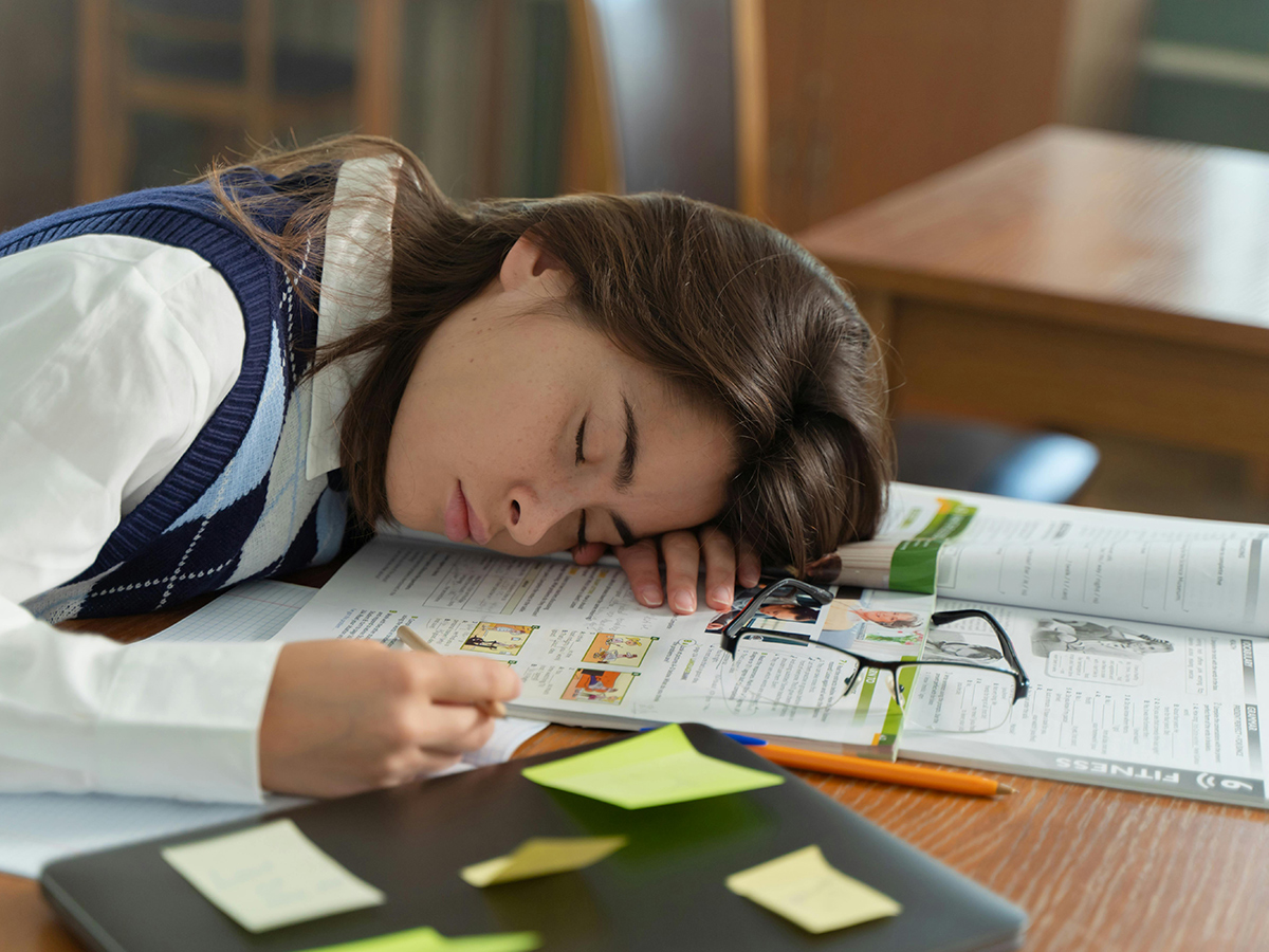 A student using an open textbook as a pillow while sleeping at a desk covered in notes and papers.
