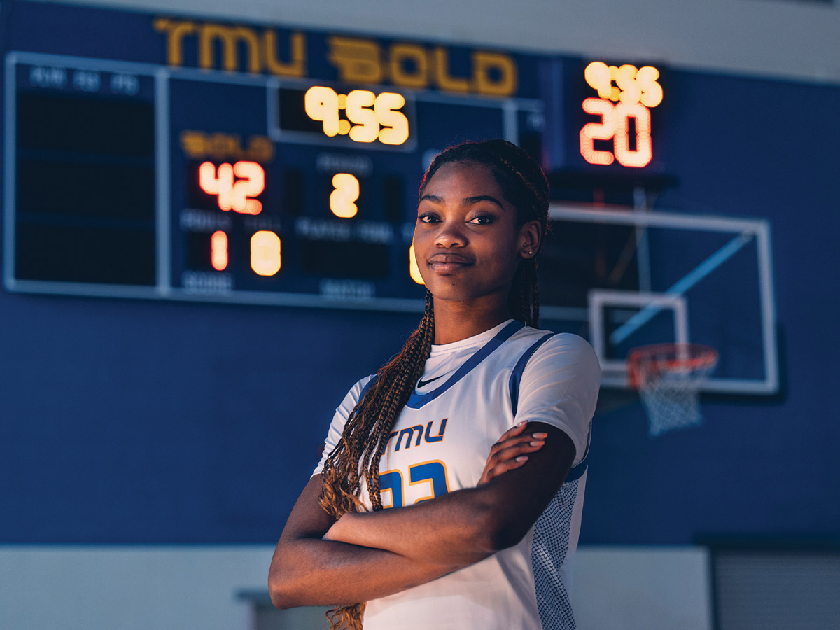 A TMU Bold athlete poses with crossed arms and a confident smile on the home basketball court.