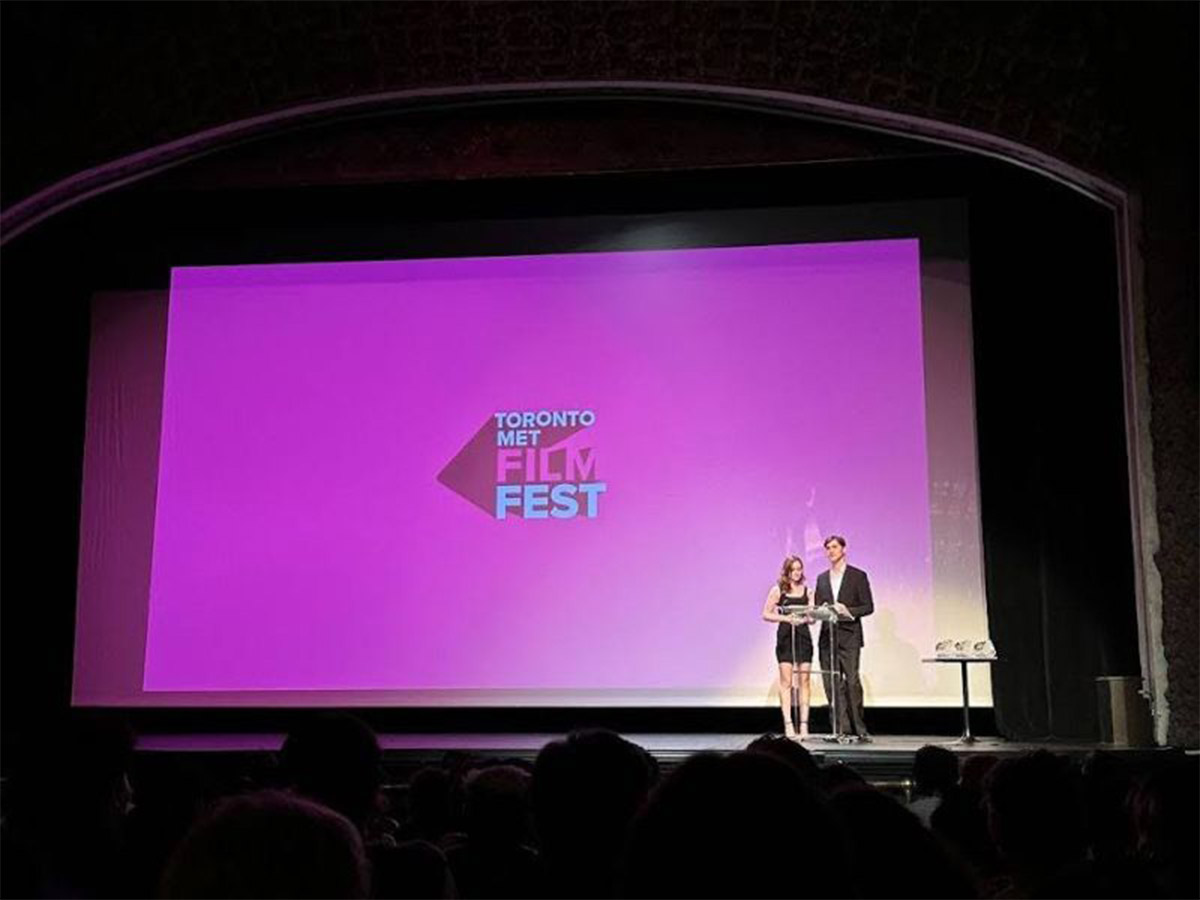 A pair of hosts speak in front of an audience at the nighttime screenings of the Toronto Met Film Festival, where fourth-year Image Arts: Film students showcase their capstone projects.