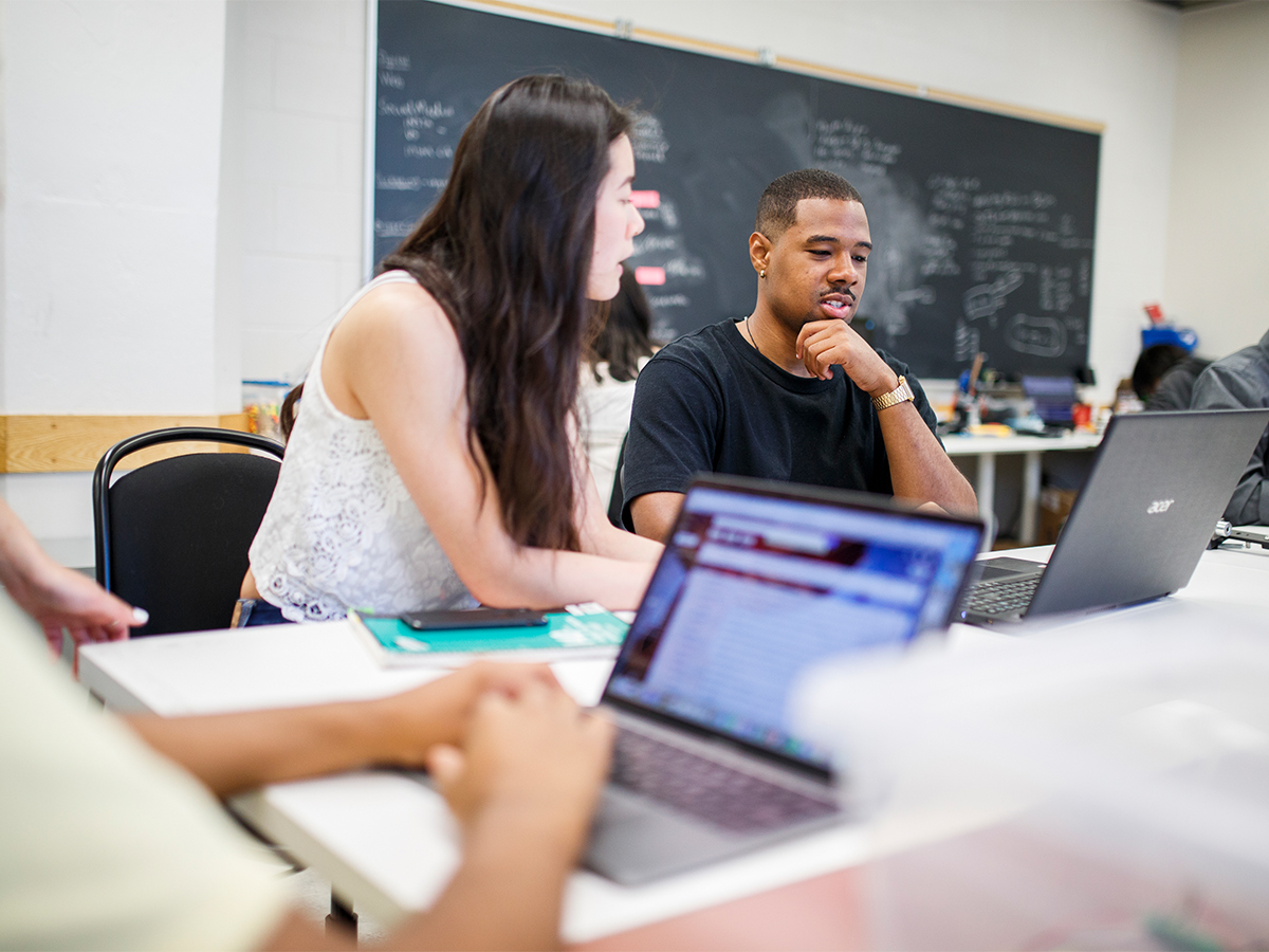A pair of students sit at a desk in a classroom and chat.
