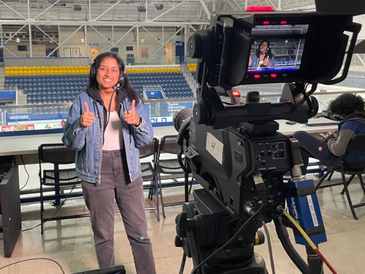 Ria gives 2 thumbs up in front of a broadcast camera at the ice rink of the Mattamy Athletic Centre.