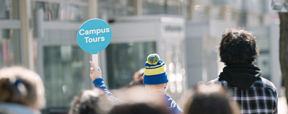 Brings you to the book your in-person campus tour section. Student guide holding a blue campus tours sign leading a group of visitors while walking past the George Vari engineering building