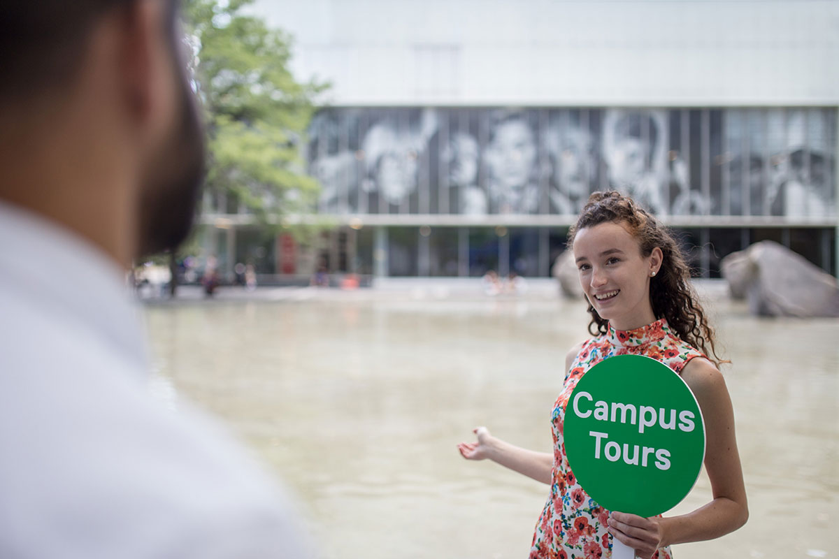 A student tour guide holding a "Campus Tours" sign gestures to Lake Devo on campus.