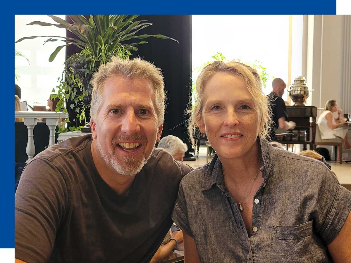 A couple enjoys beverages at a bar, against a backdrop of tall, sunlit windows, large plants and a high chandelier.