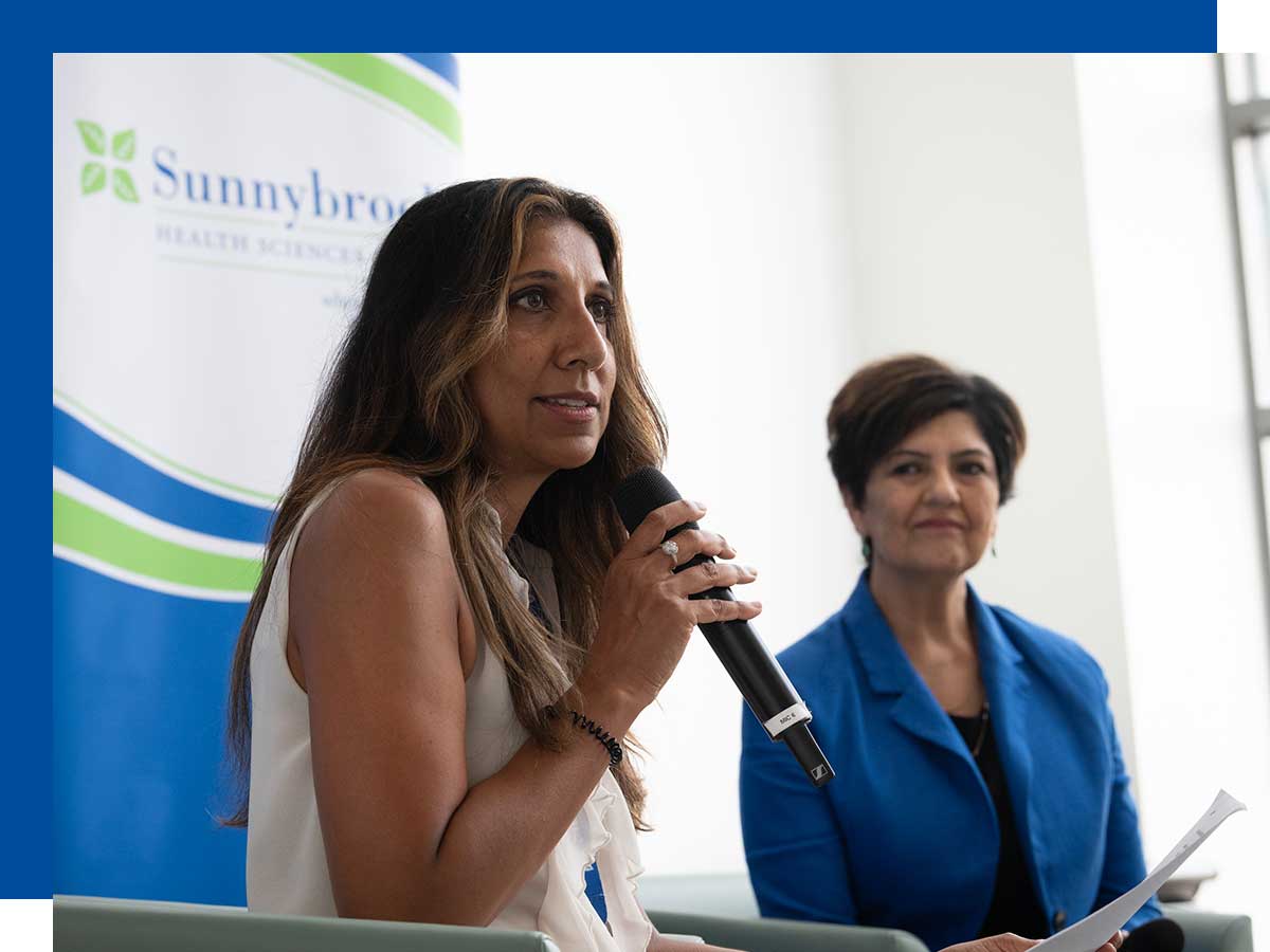Taggar holds a microphone while another woman looks on; background signage says Sunnybrook Health Sciences