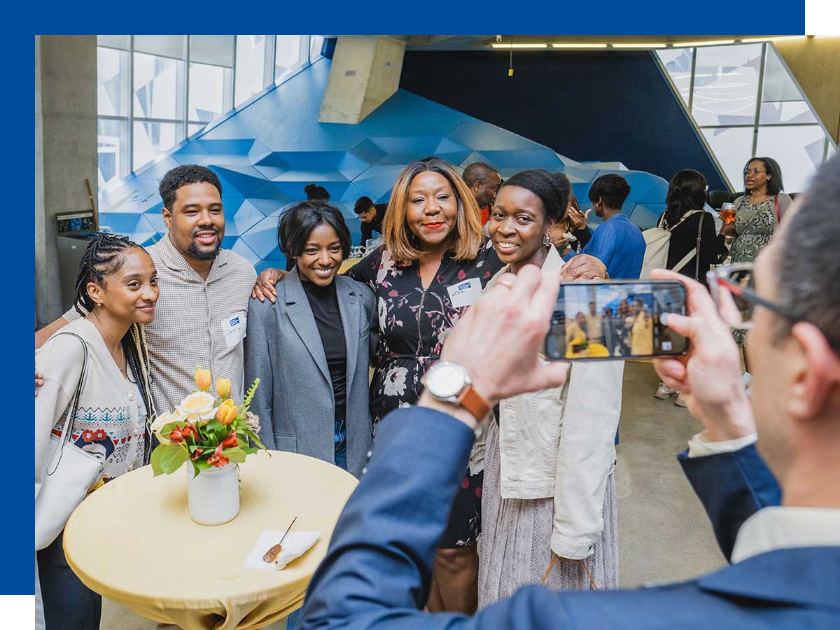5 people posing for a group photo and a man taking the photo on his phone