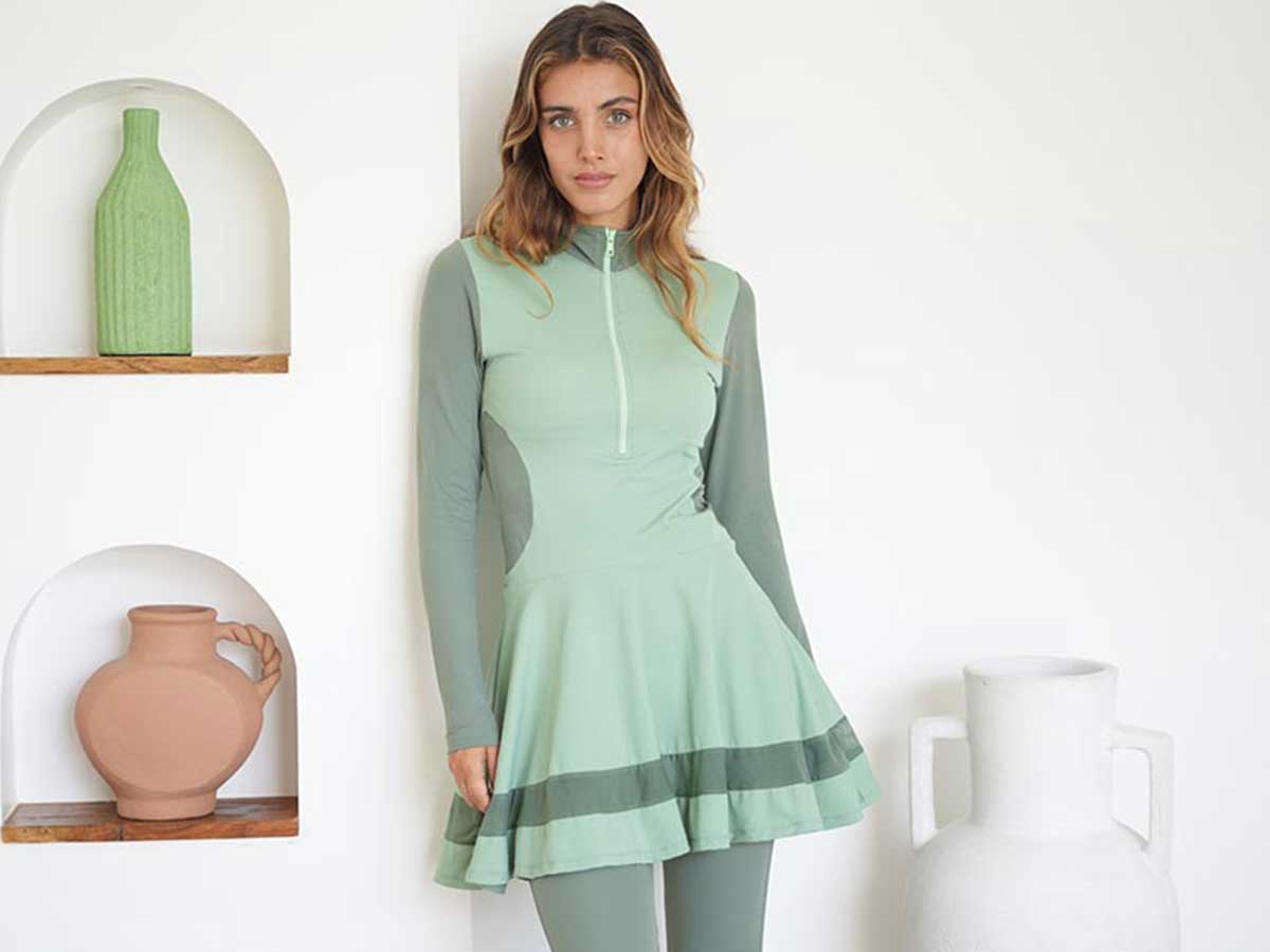 Young woman wears a light green full-coverage swimsuit as she leans against a wall shelf with colourful vases