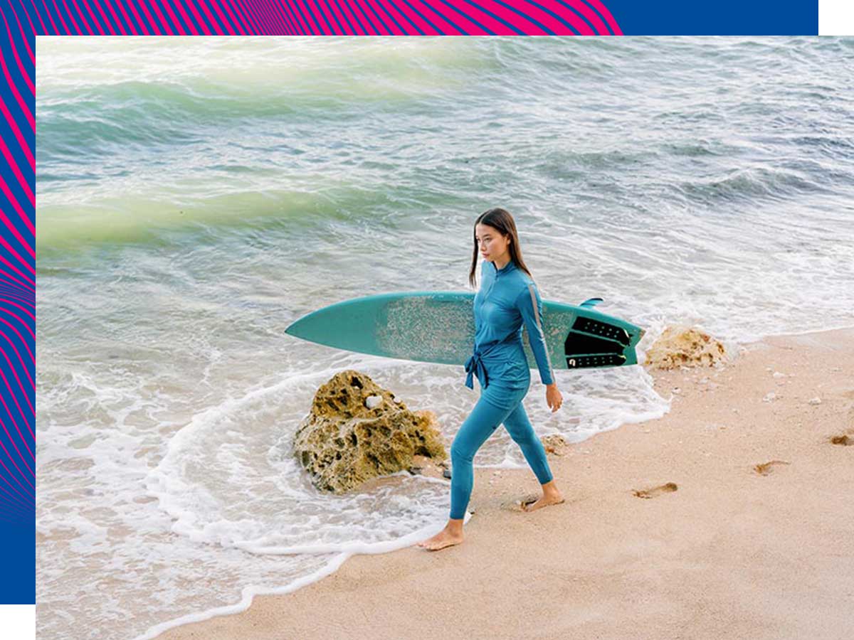 Young woman wearing skirted rash guard and leggings walking along a beach an holding a surfboard