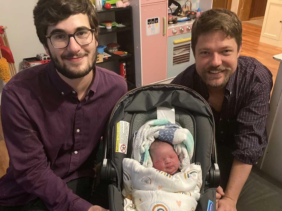 Two men kneel on the floor, between them is a newborn asleep in a baby carrier