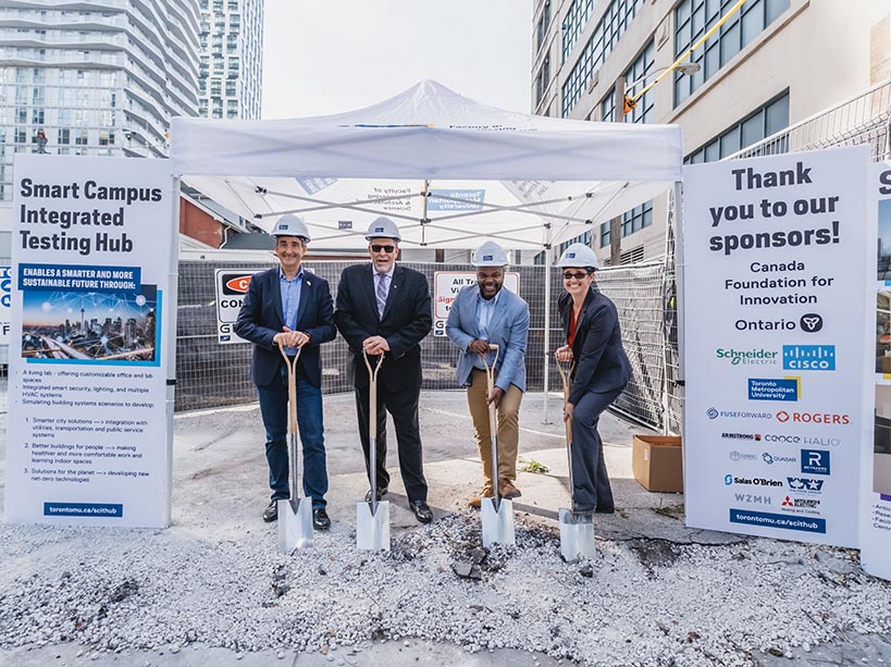 Four people wear hard hats and hold shovels on a construction site.