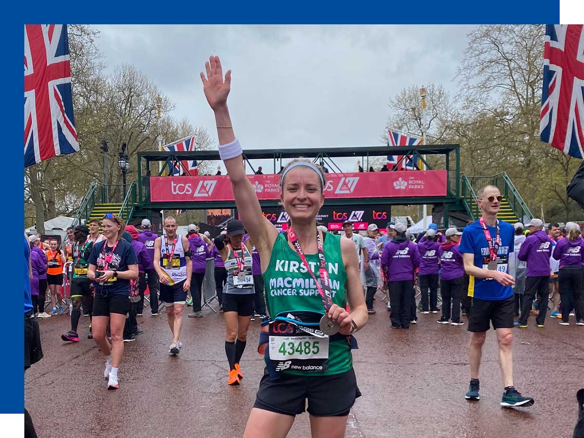 A woman poses with one arm in the air and the other hand holding a medal around her neck at the finish line of a race with other runners in the background. 