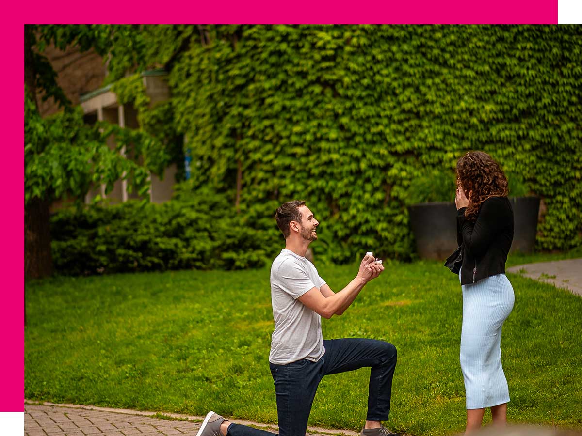 With an ivy-covered building behind them, a man on bended knee presents an engagement ring to a surprised woman.