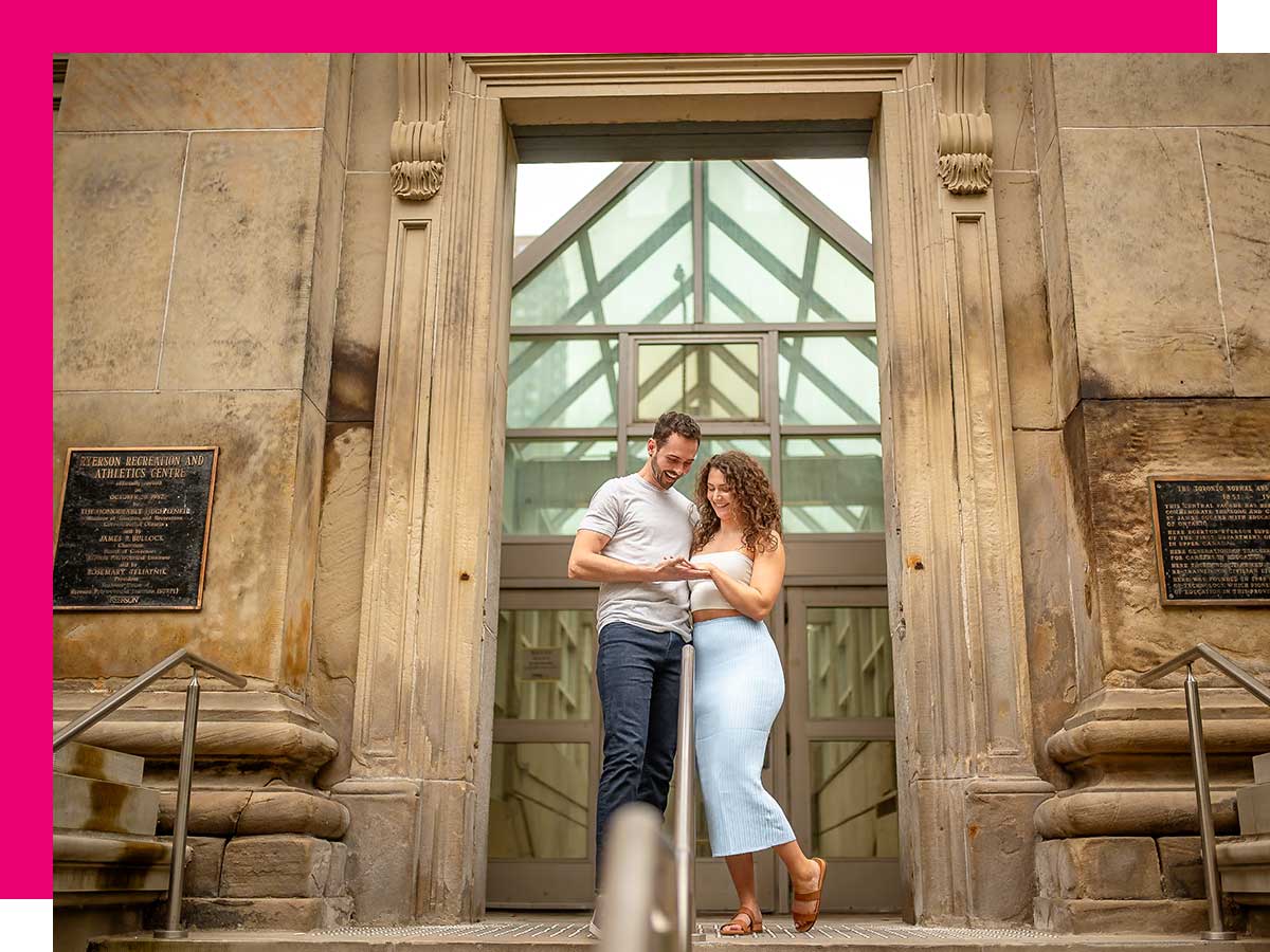 A man and woman stand at the top of a set of stairs leading to a building entrance, her left hand held in his right hand, together gazing at her engagement ring.