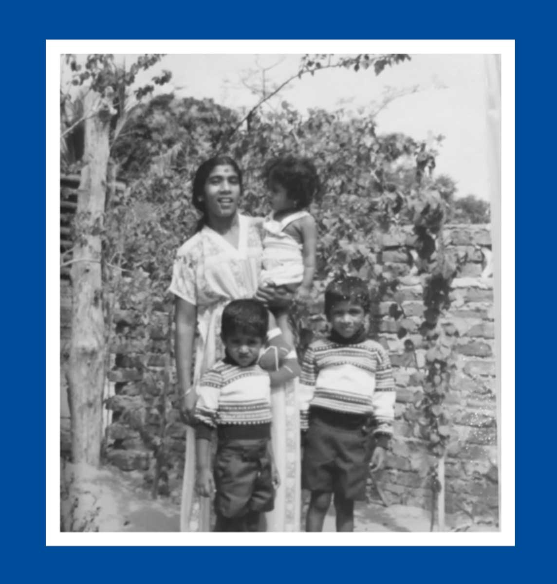 Black and white photo of a woman holding a little girl in her arms. Two boys stand in front, wearing matching sweaters and shorts.