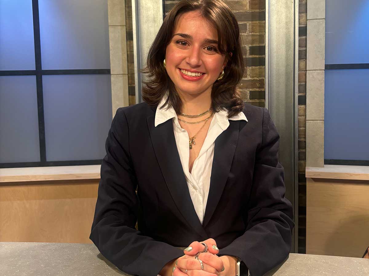 Woman smiles and sits behind broadcast news desk.
