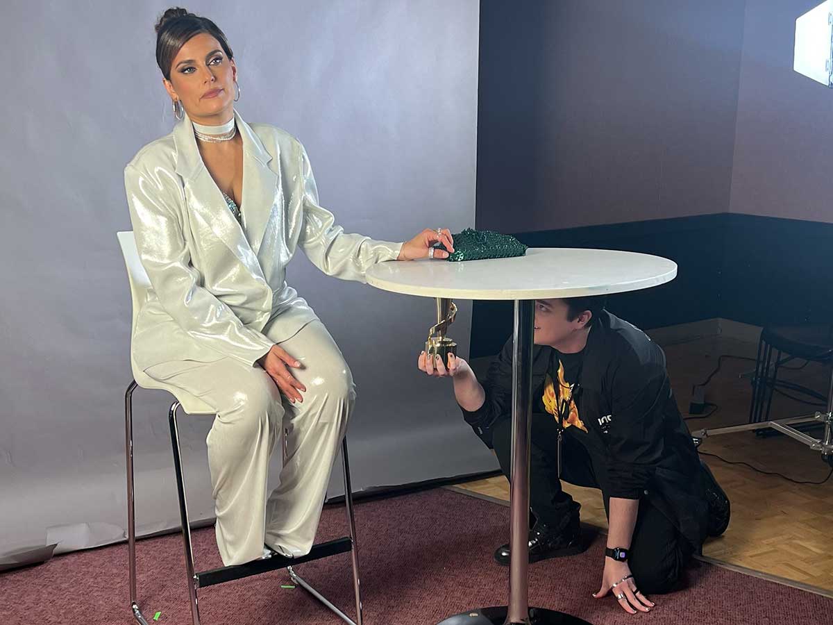 A studio with white screen backdrop, mic and camera lights. Woman dressed in white sits at a high table while man kneels on floor, checking set-up