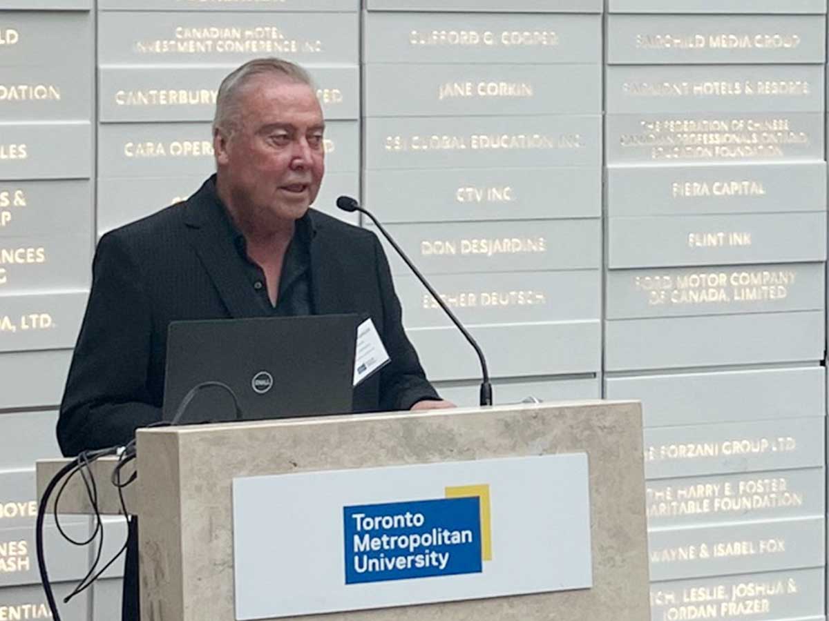 Man stands at podium, a backdrop of names on plaques behind him.