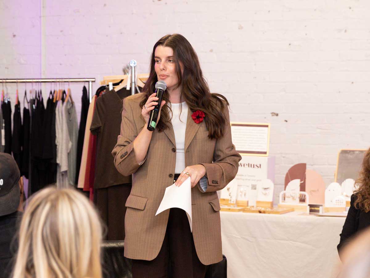 A woman holds a mic as she address a crowd in front of a rack of clothing.