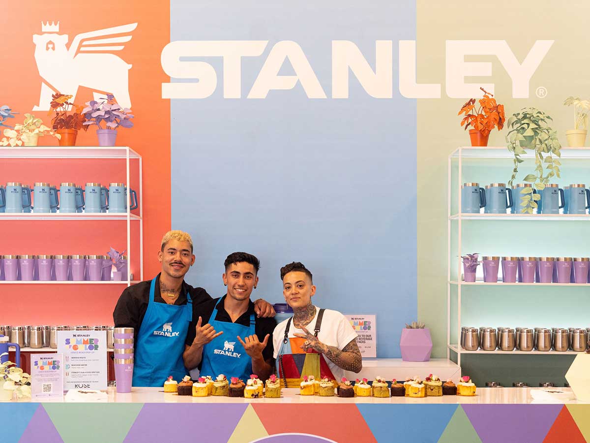 Three people wearing aprons stand behind a counter with cupcakes in front of a backdrop for Stanley. 