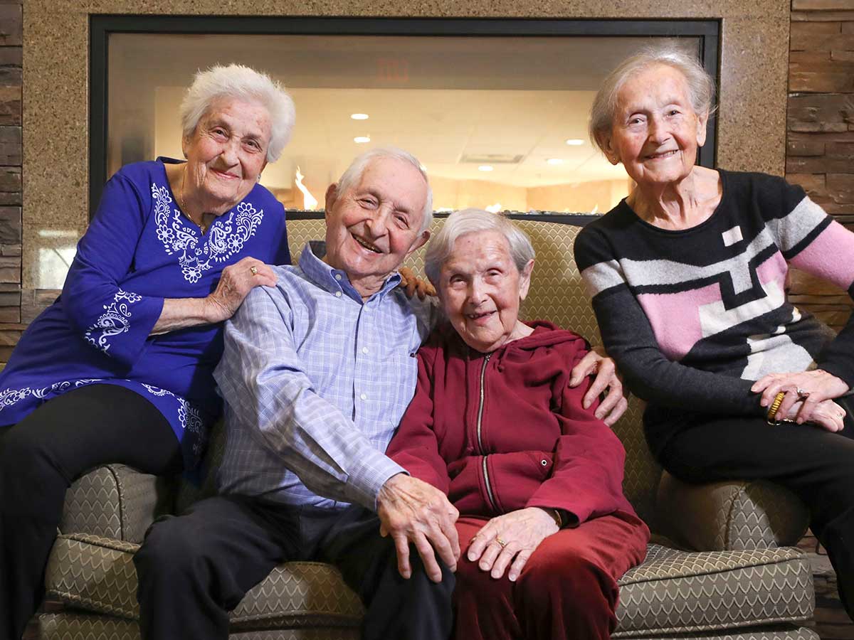 All smiling, a man and woman seated on a couch, as two women seated on the couch arms lean in, a lit fireplace and wood and brick walls behind them.