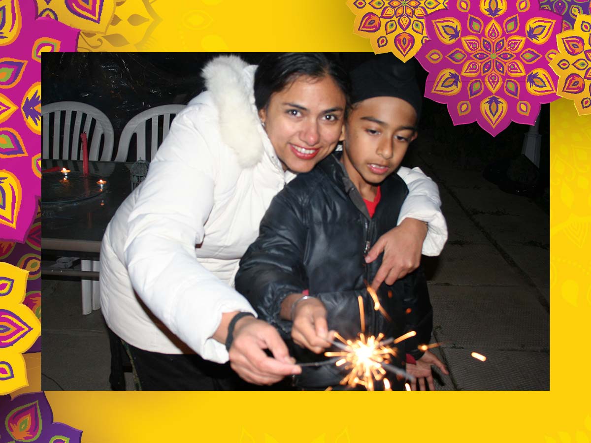 Standing outside at night, two people in winter coats hold sparklers that starburst with light. A table behind them holds lit tea lights.
