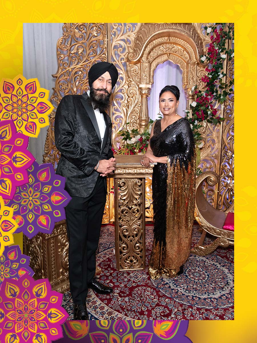 A man and woman dressed in formal attire stand at a backdrop decorated for Diwali.