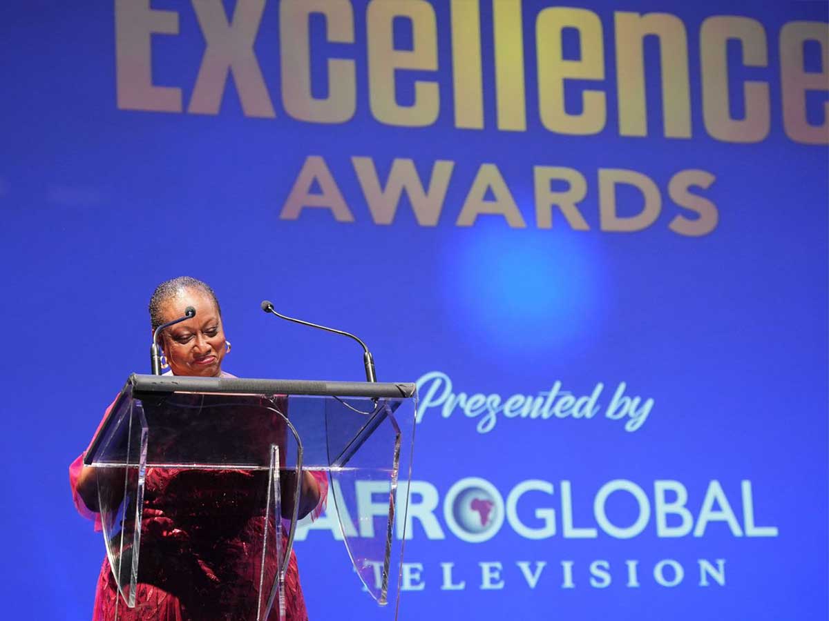 A woman in a red dress stands in front of a podium while giving a speech.