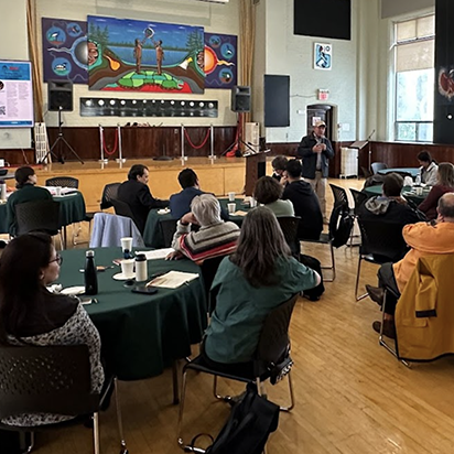A large group of people are seated at round tables with green cloths, facing a stage. On the stage, a man in a blazer is speaking into a microphone, and behind him is a large, colorful mural depicting two people by a lake with a forest in the background.