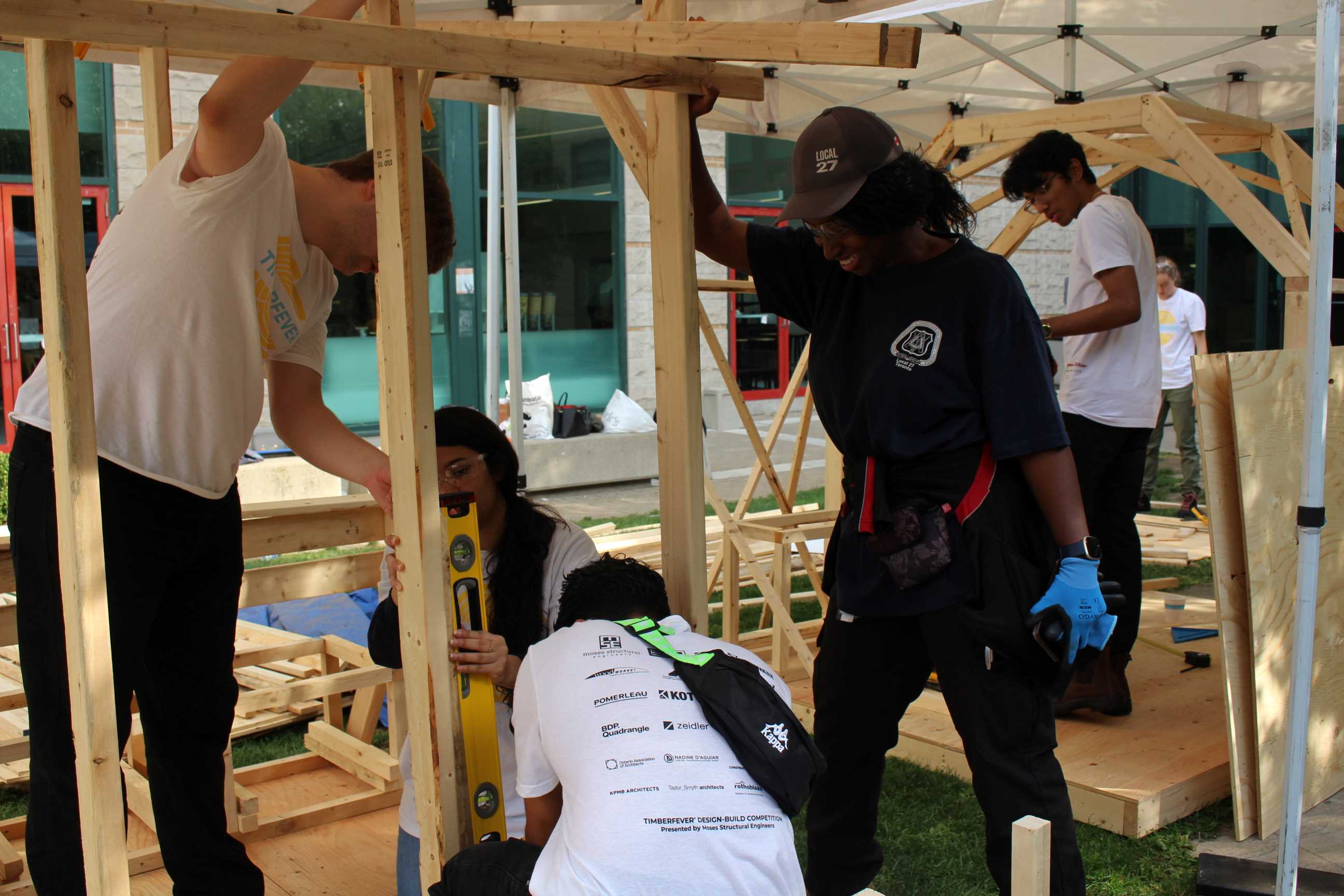 Students measuring wooden planks while a carpenter mentor holds up a plank. 