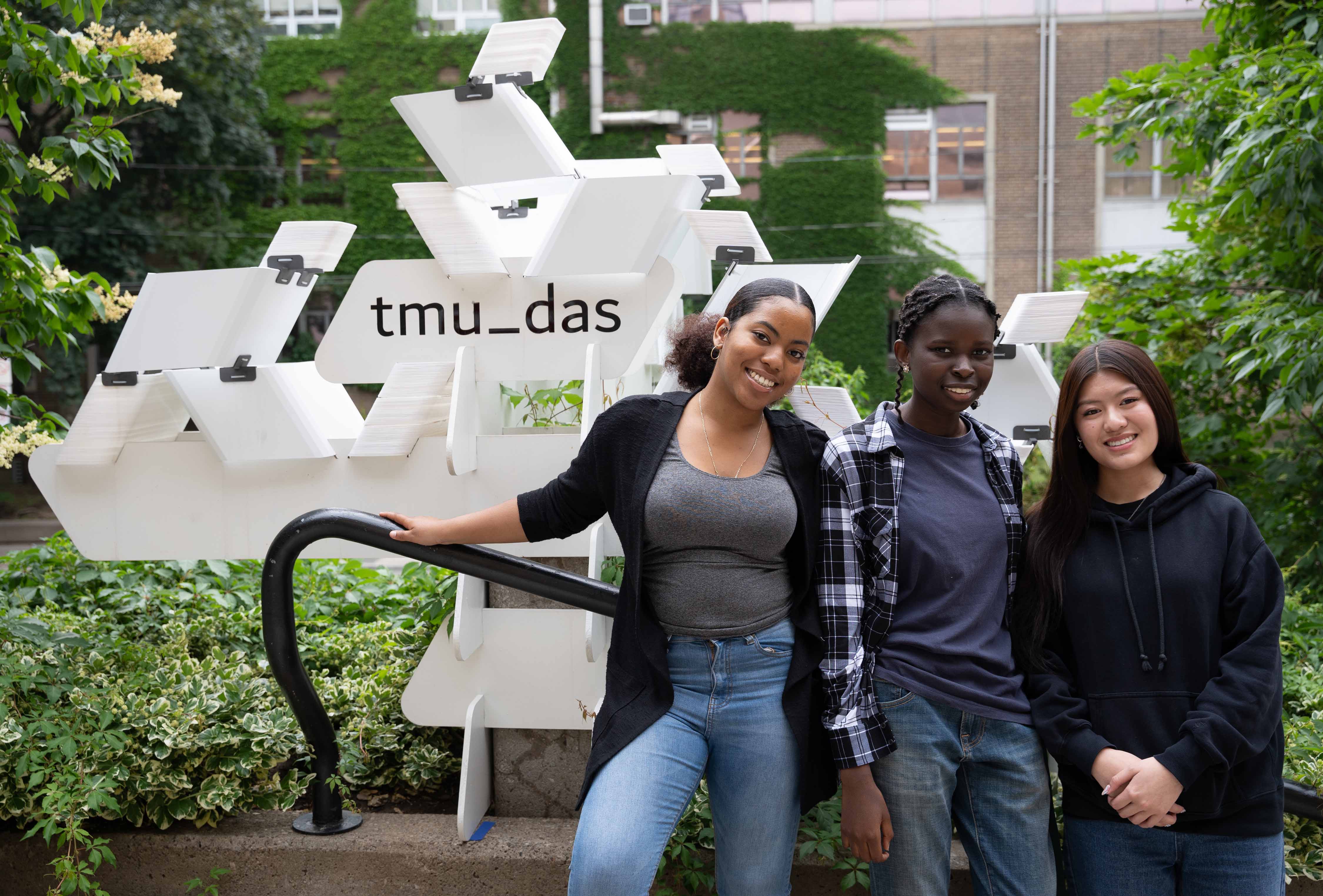 Jasmin Tran, Ana Marya Queralta Pantoja, and Rita Mouping smiling in front of the Cloud structure on the entry stairs of the Architecture Building
