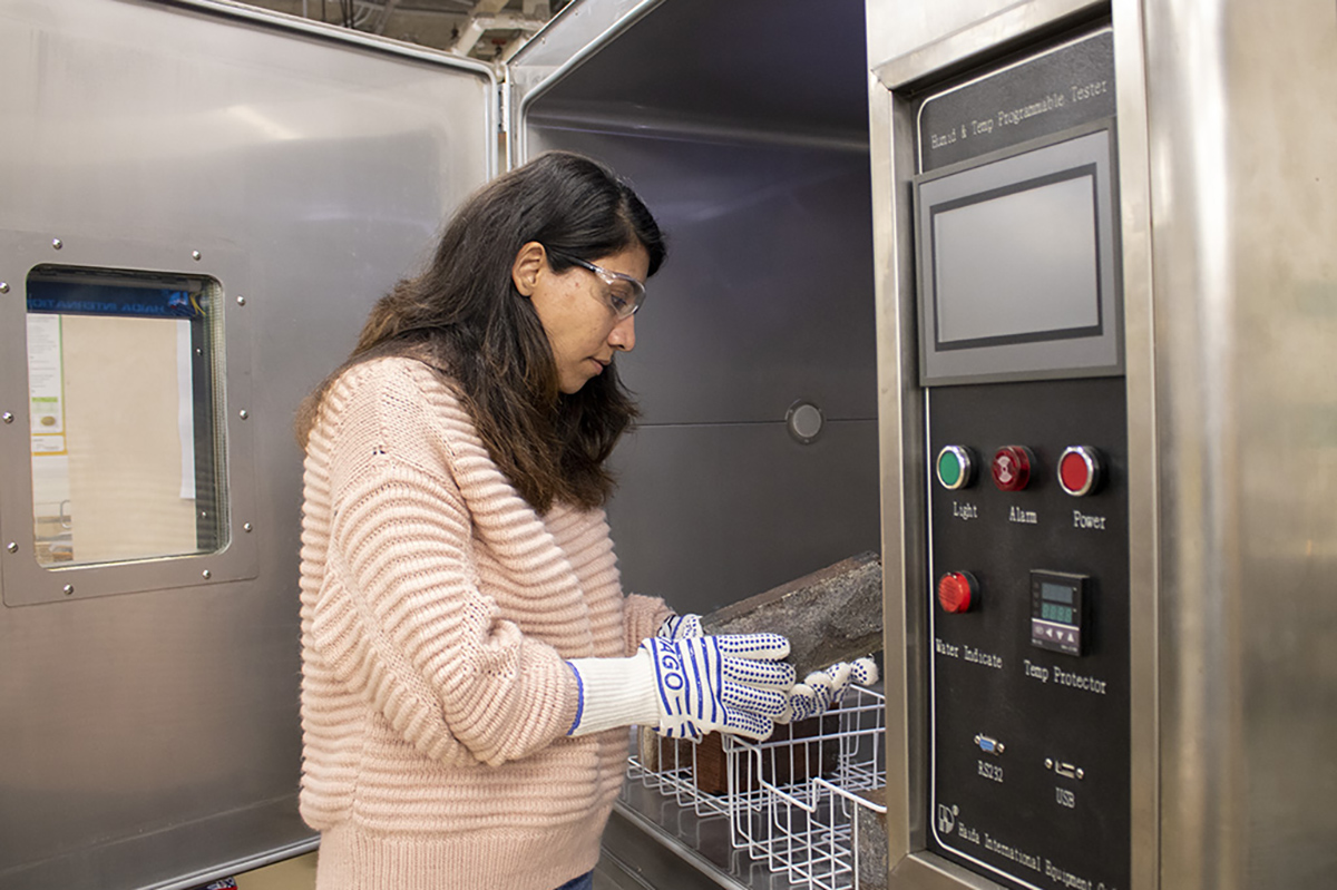 In the Building Science Lab, a student wearing safety glasses and protective gloves holds a block of building material inside a temperature testing machine. 