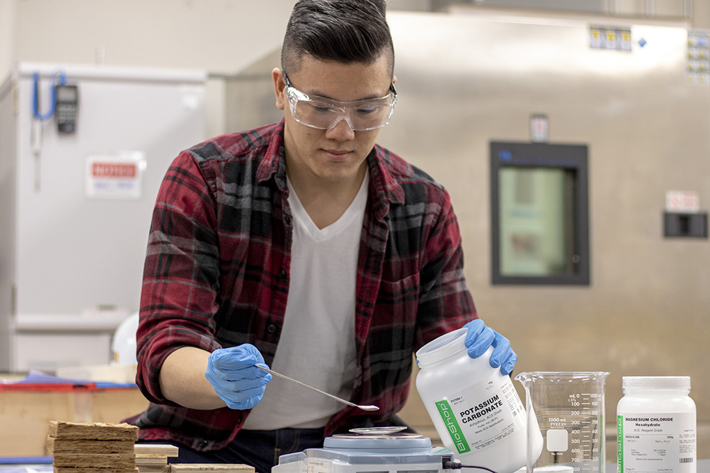 A student wearing safety glasses and protective gloves weighs a white powder on a scale. 