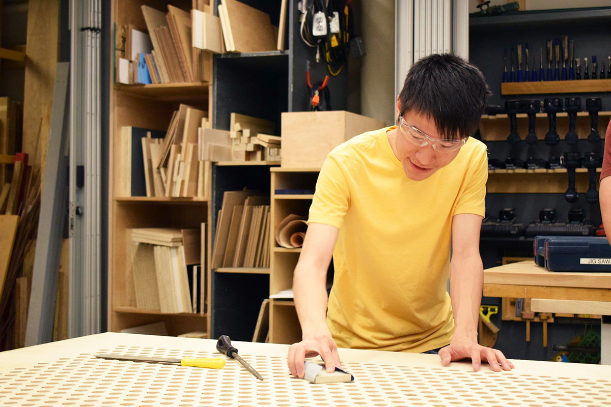 In the Bench Room, a student wearing safety glasses sands a large piece of perforated wood.