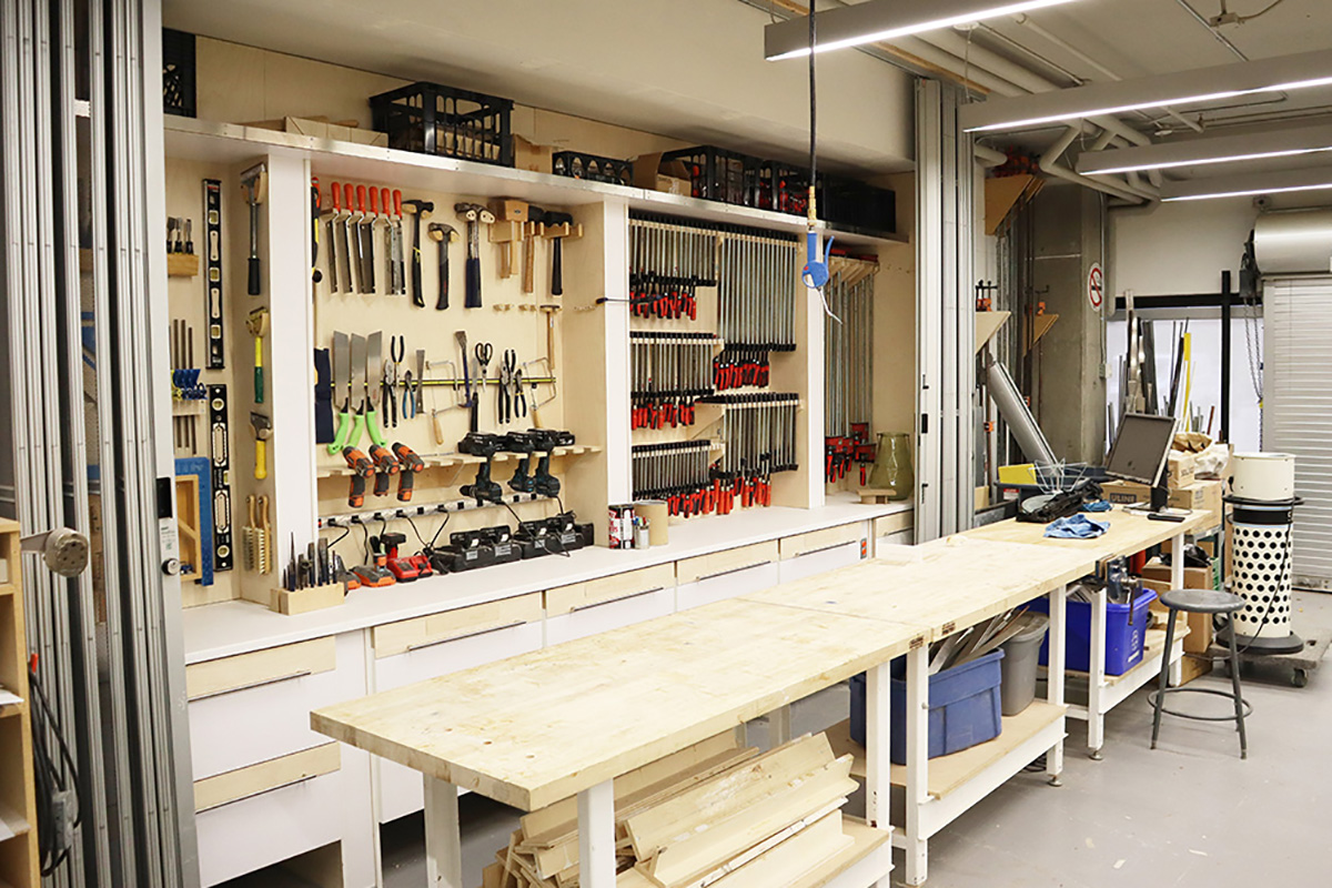  shelving unit stores a variety of tools, including electric drills, levels and hammers. Three worktables are located in front of the shelving unit. 