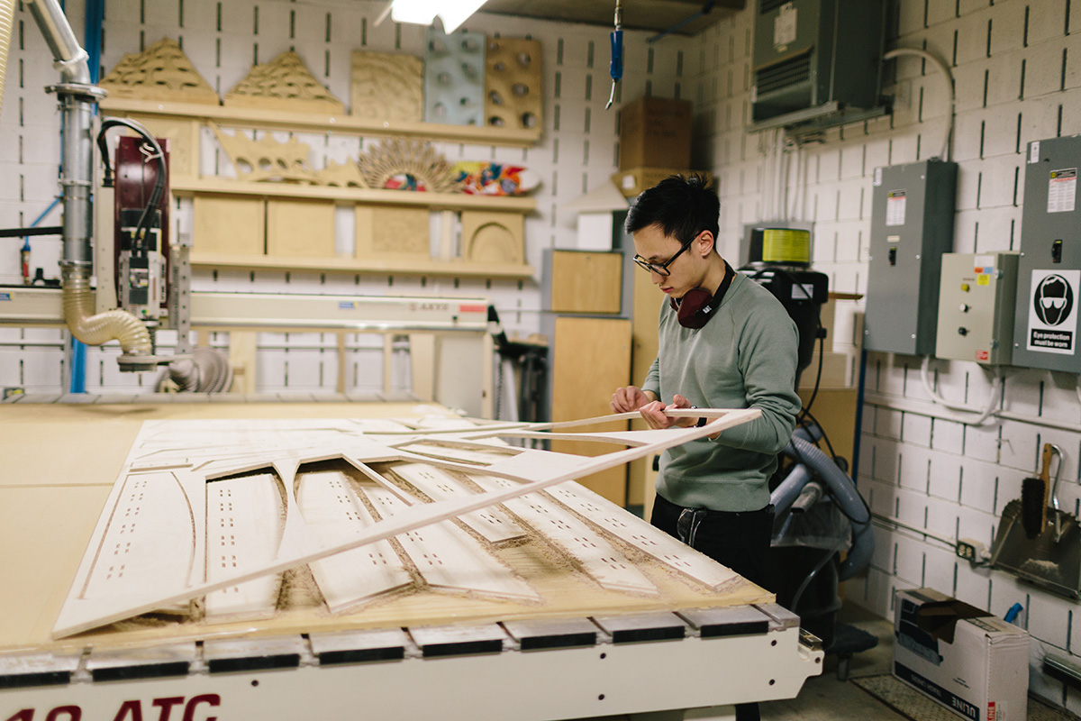 In the Robot and Computer Numerical Control Lab, a student examines pieces of intricately cut wood, which are laid out on a large work table. 