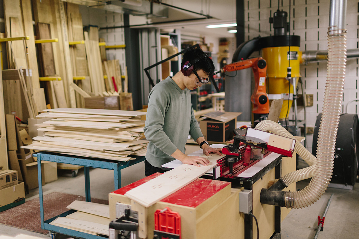 A student wearing safety glasses and ear protection cuts a piece of wood using a CNC machine.