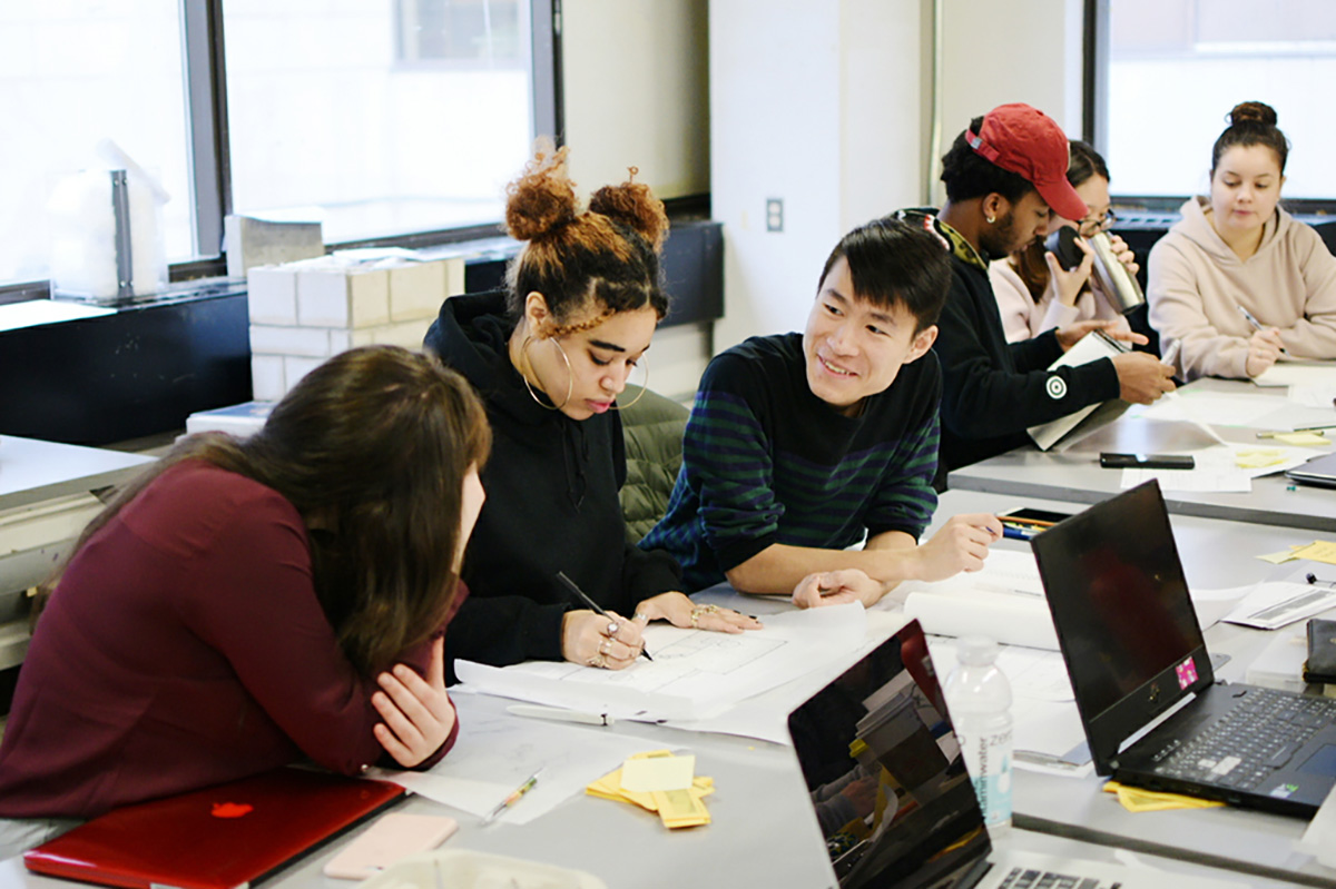 Two small groups of students work together at a communal desk covered in papers. 
