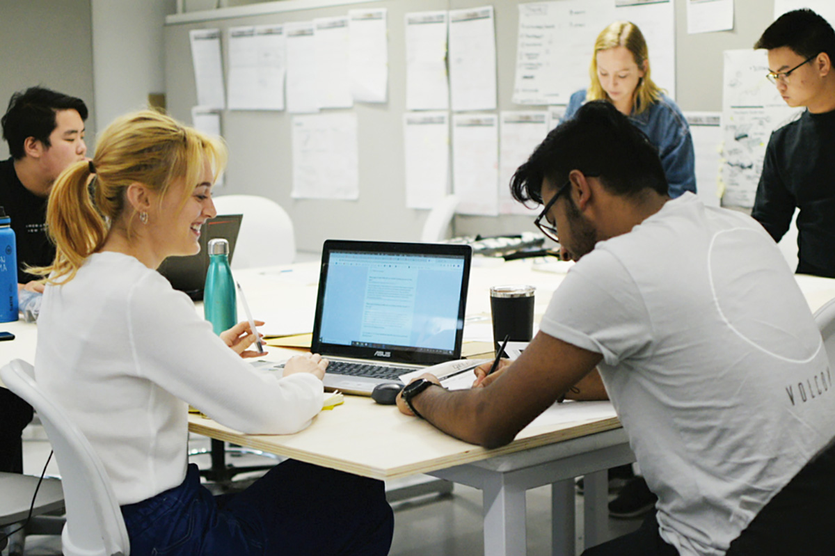 In their studio space, five students work around a large desk covered in papers and laptops. Diagrams and other papers are pinned to a wall. 