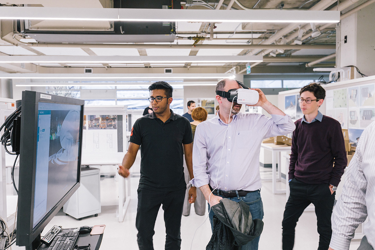 A faculty member wears a virtual reality headset while two students present work on a large screen. 