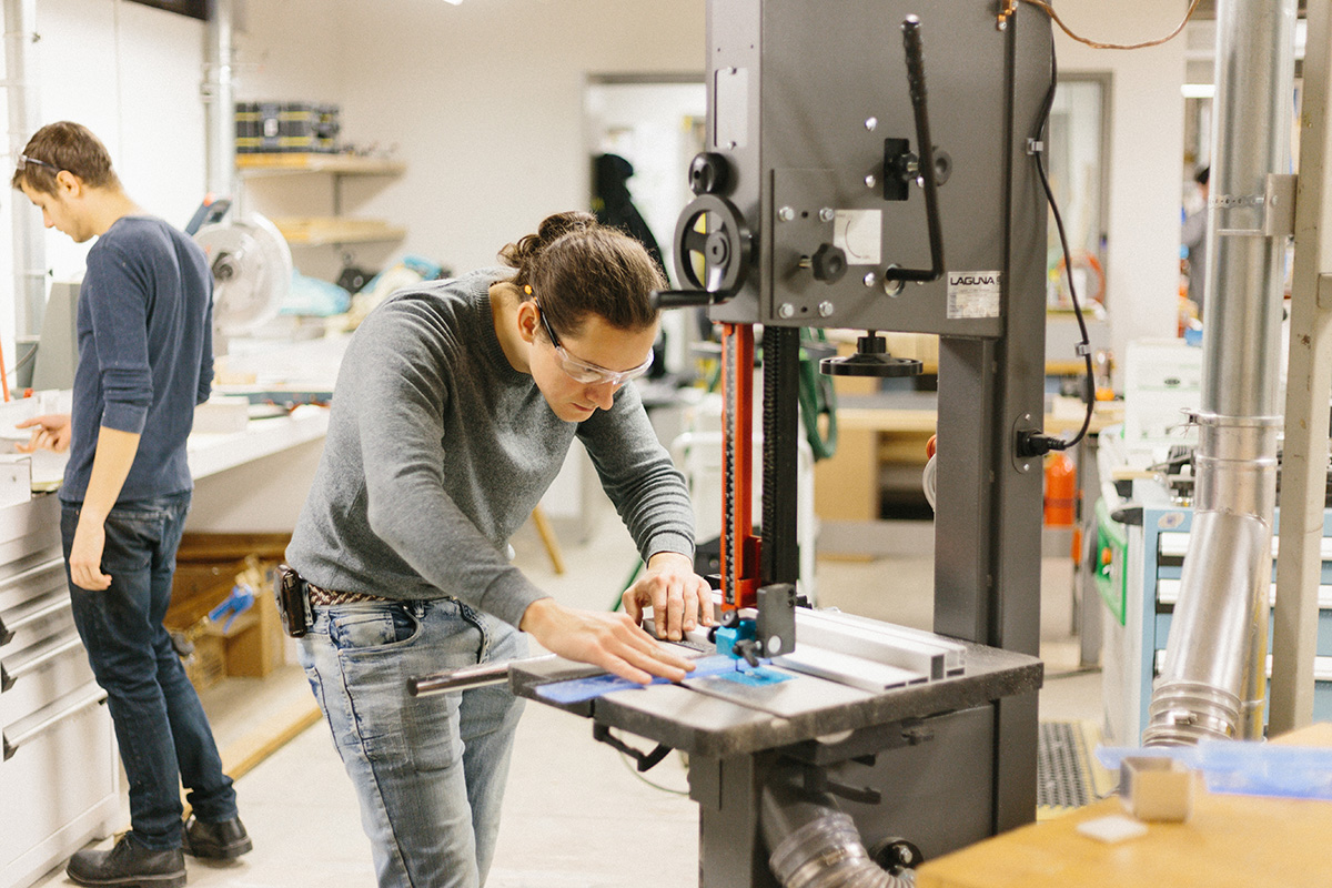 In the Woodshop, a student wearing safety glasses carefully cuts a piece of blue material using a vertical band saw. 