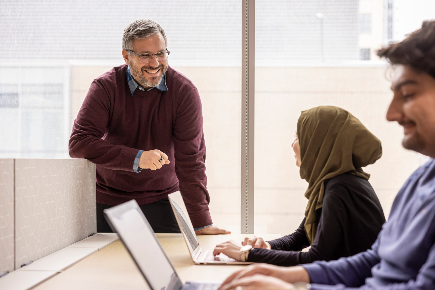 A professor smiles at a student working at a table sitting beside another student. Both are using laptops