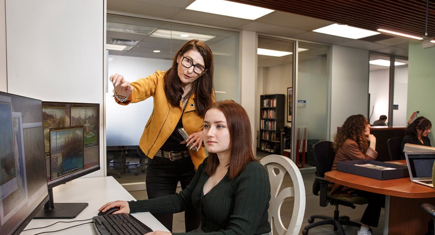 Three students look at one laptop, one points at the screen.