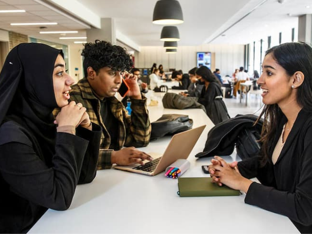 Three students sit at a white table in a modern study space, two in conversation, one using a cellphone 
