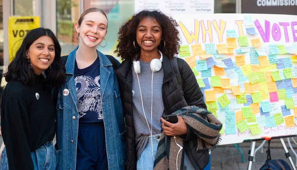 Three students smiling standing infront of a poster "Why I vote"